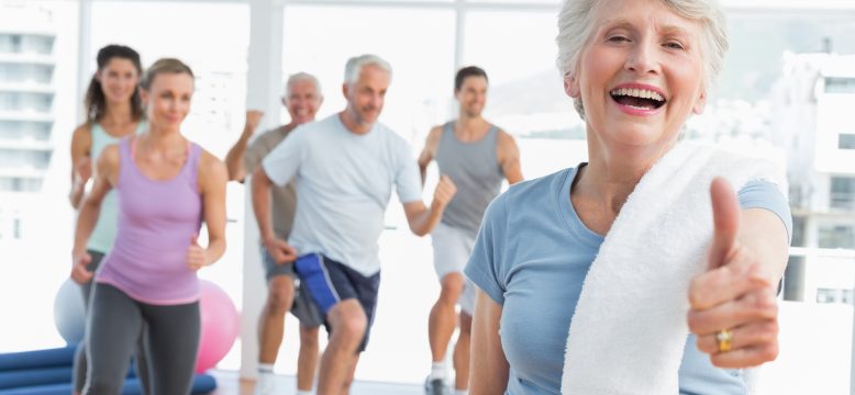 How to stay pain free and have happy, healthy feet for longer! Cheerful senior woman gesturing thumbs up with people exercising in the background at fitness studio