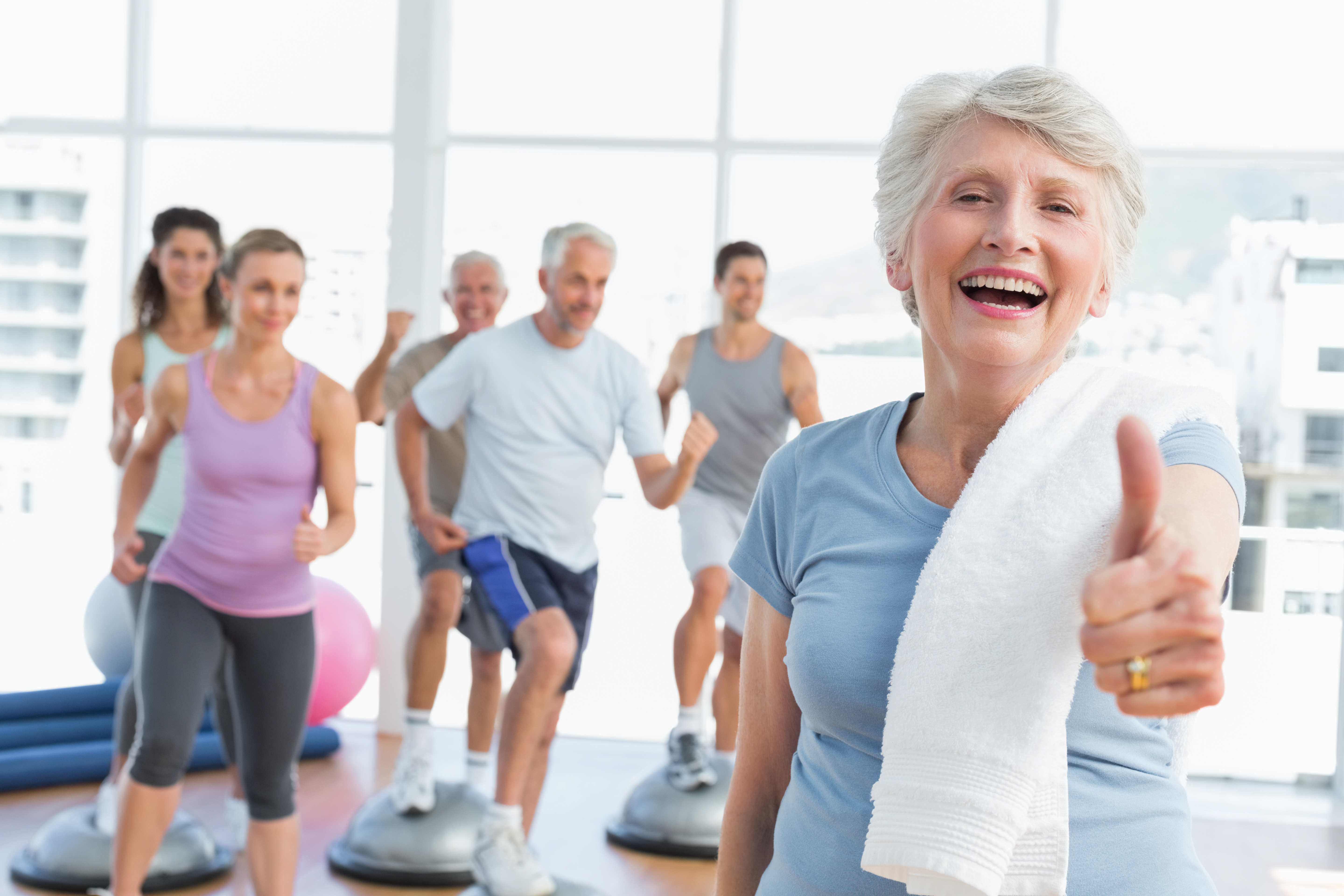 Cheerful senior woman gesturing thumbs up with people exercising in the  background at fitness studio - Foot & Leg Centre